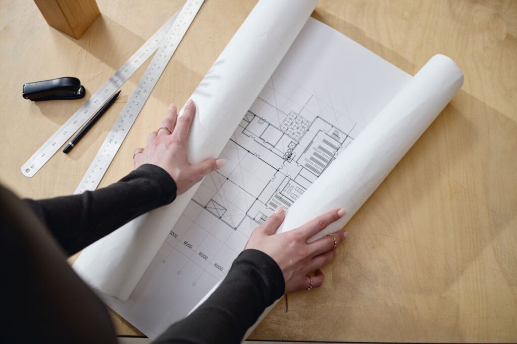 Caucasian young adult woman unrolling architectural blueprint on wooden desk, hands visible arranging technical drawing, metal ruler and stapler placed nearby, working on project 